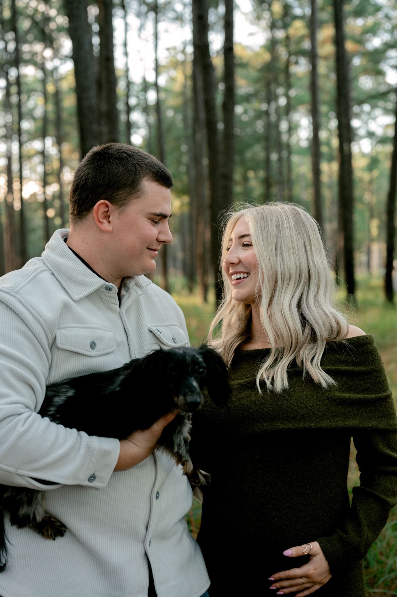 Husband and wife holding Dachshund beneath pine trees.