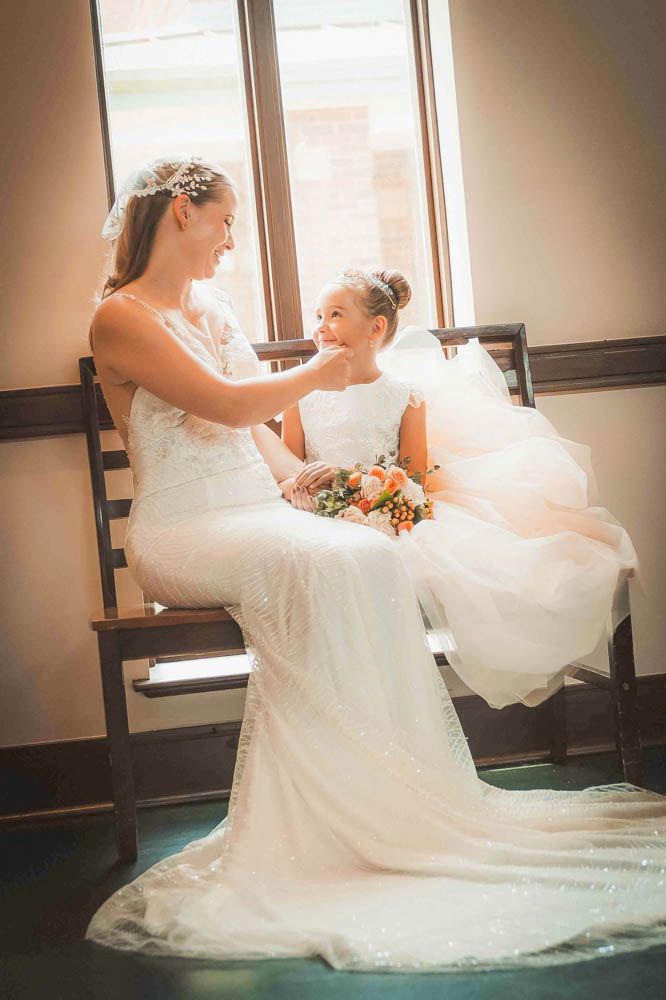 Bride in white gown gently touches flower girl's face while seated together on a bench near a sunlit window.