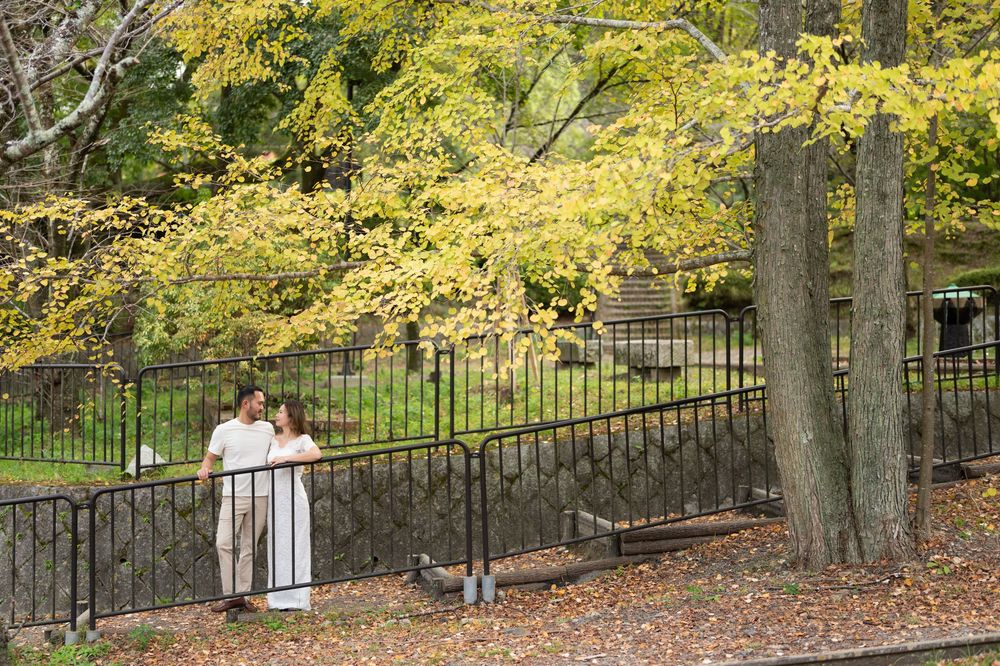 Couple in casual white outfits embracing beside a black iron fence under golden autumn trees with fallen leaves in a park.