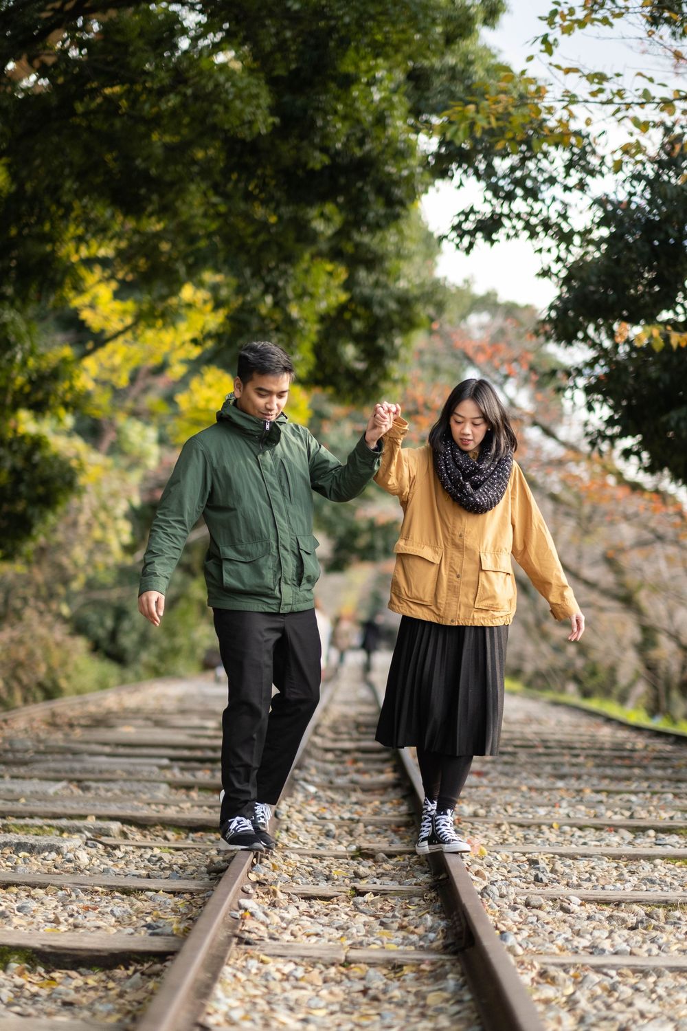 A couple holds hands while walking along old railroad tracks surrounded by colorful autumn trees on a sunny day.