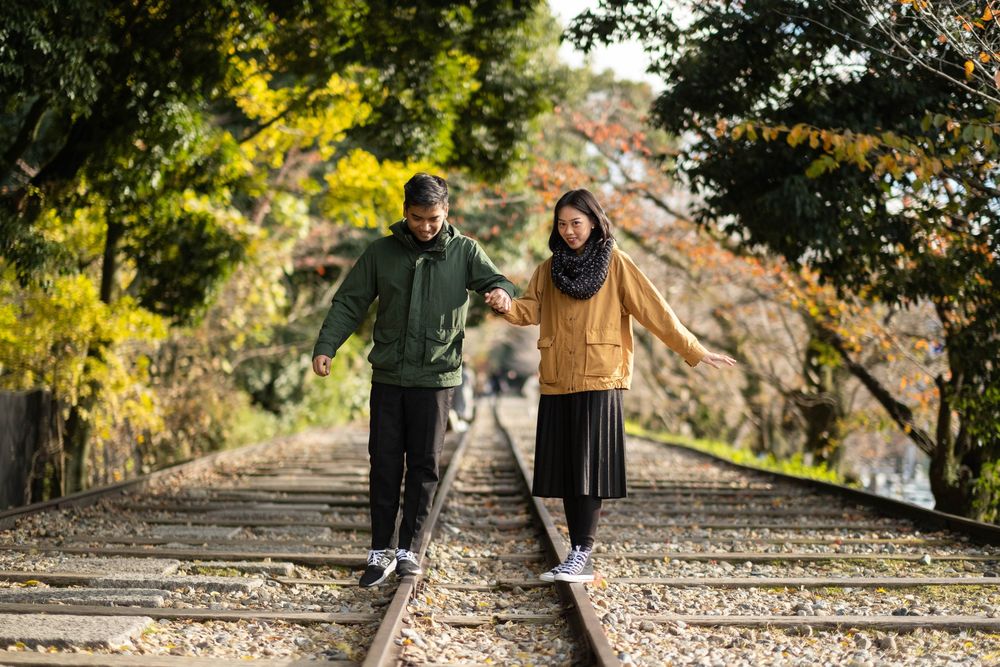 Couple holding hands while balancing on separate rails of an old railroad track surrounded by colorful autumn trees.