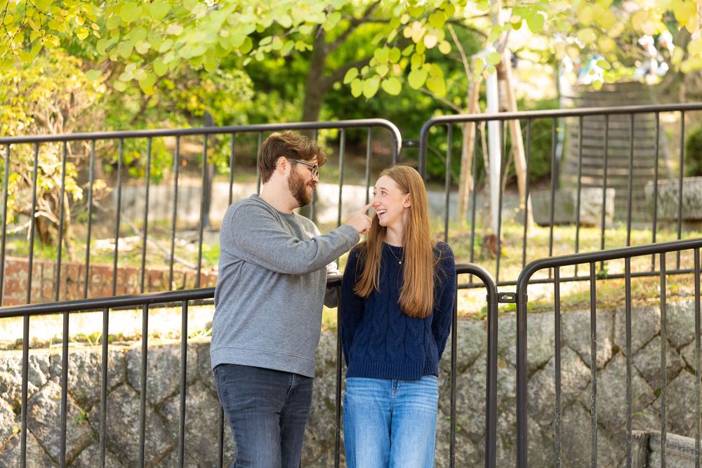 A bearded man in a gray sweater and a woman with long hair in a navy top smile and talk beside a black metal fence outdoors.