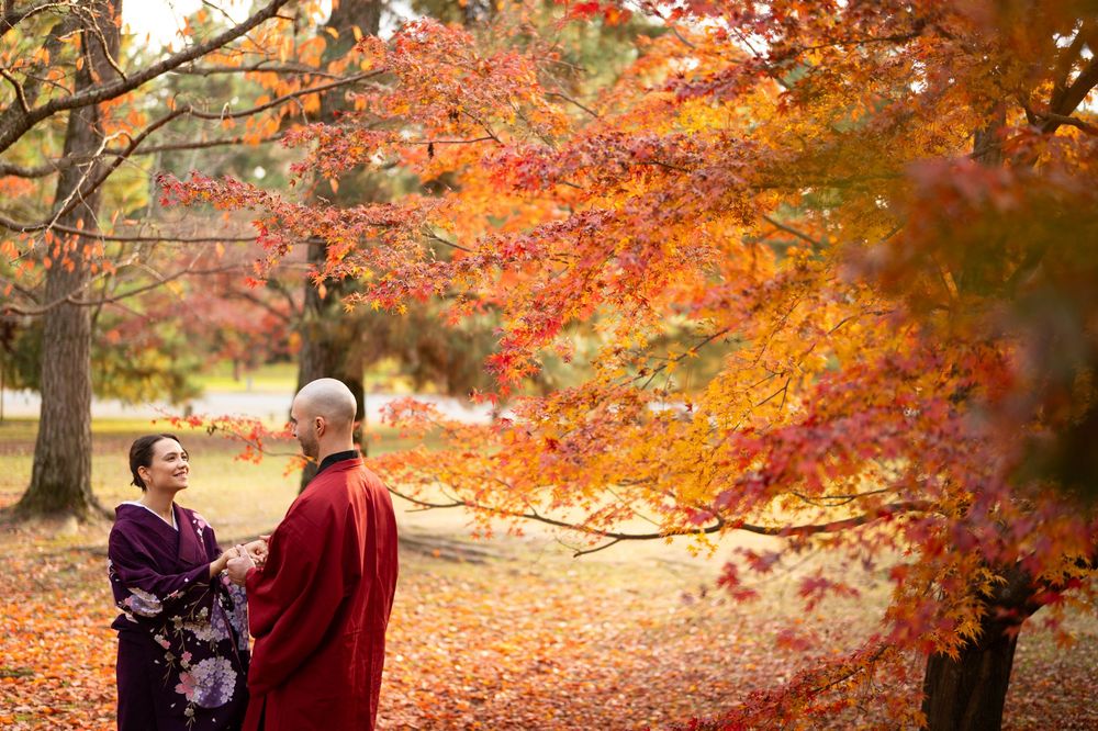 Couple in traditional Japanese kimono standing together beneath vibrant red and orange autumn maple trees in a peaceful park.