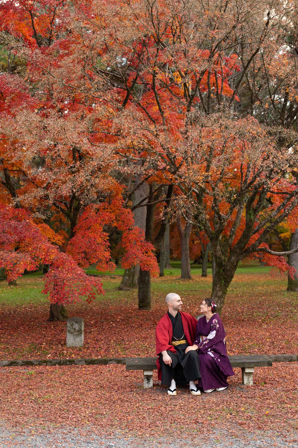 Couple in traditional Japanese kimono sit on a wooden bench beneath vibrant red autumn maple trees in a leaf-covered Kyoto park.