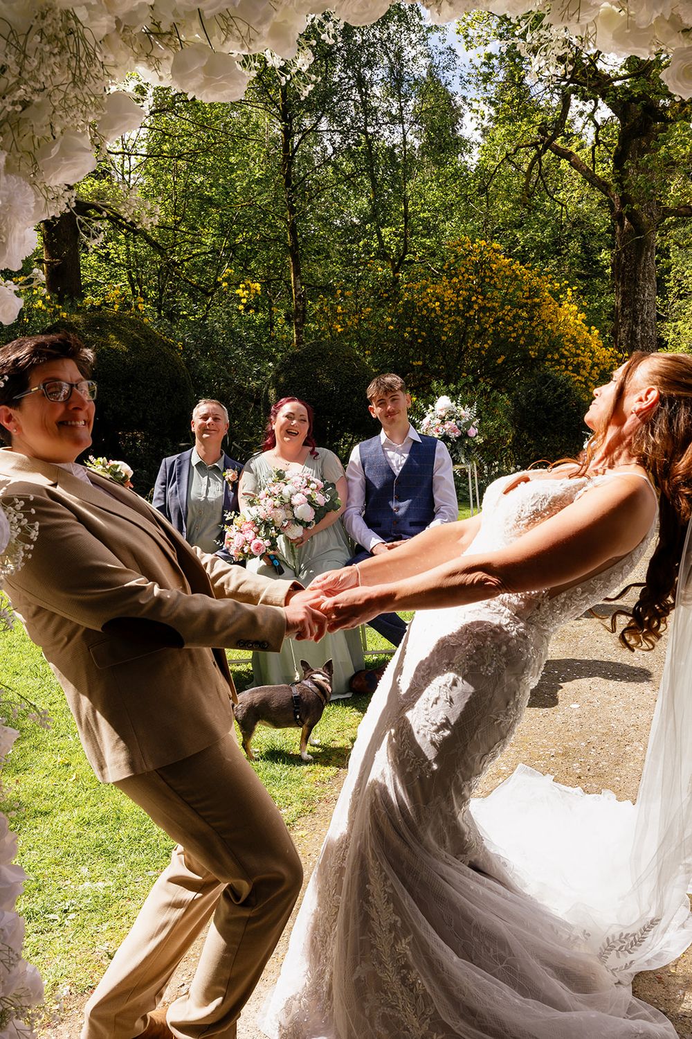 Joyful couple holding hands and leaning back during outdoor wedding ceremony with guests and floral arch.