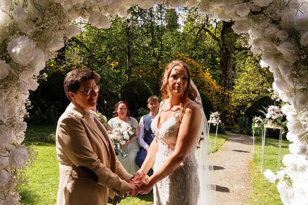 Two brides hold hands under a white floral arch during an outdoor wedding ceremony on a sunny day.