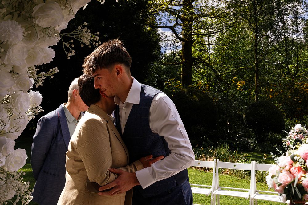 Couple sharing first kiss at outdoor wedding ceremony with floral arch and lush green trees in background.