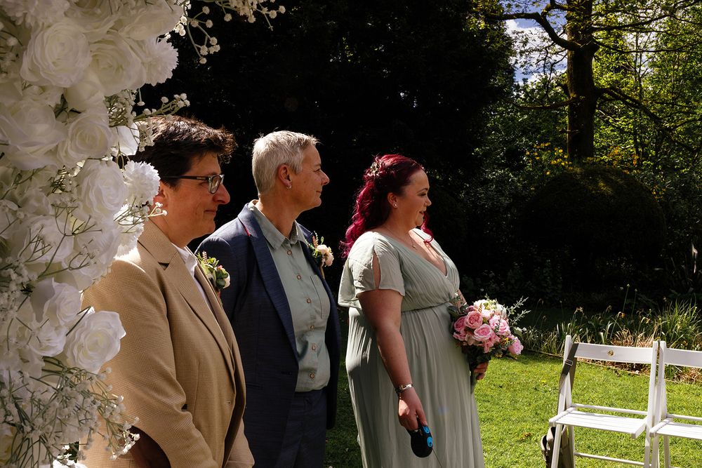 Three people standing outdoors at a garden wedding ceremony, with white floral decorations and white chairs visible.