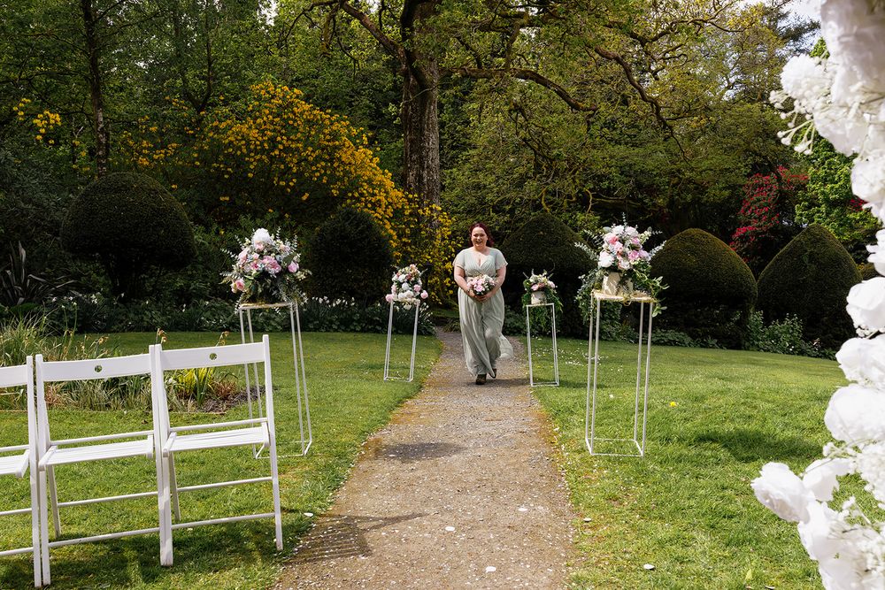 Outdoor wedding ceremony aisle with white chairs, floral arrangements on stands, and a woman walking through a lush garden.