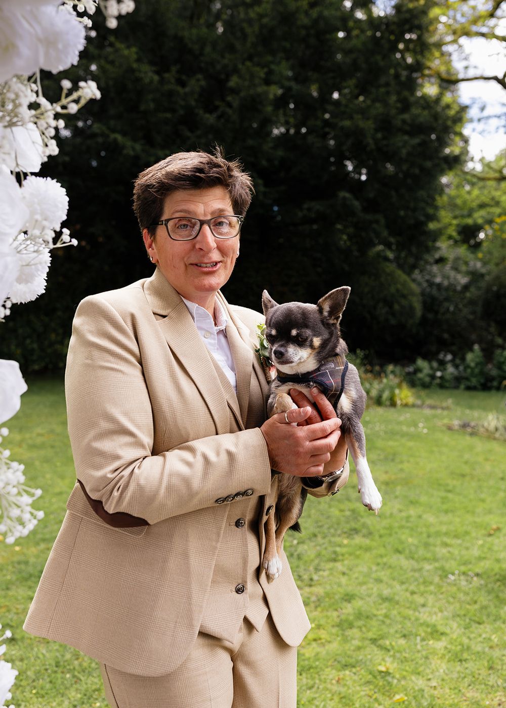 Person in beige suit holding a small Chihuahua dog at an outdoor garden wedding ceremony with white floral decorations.