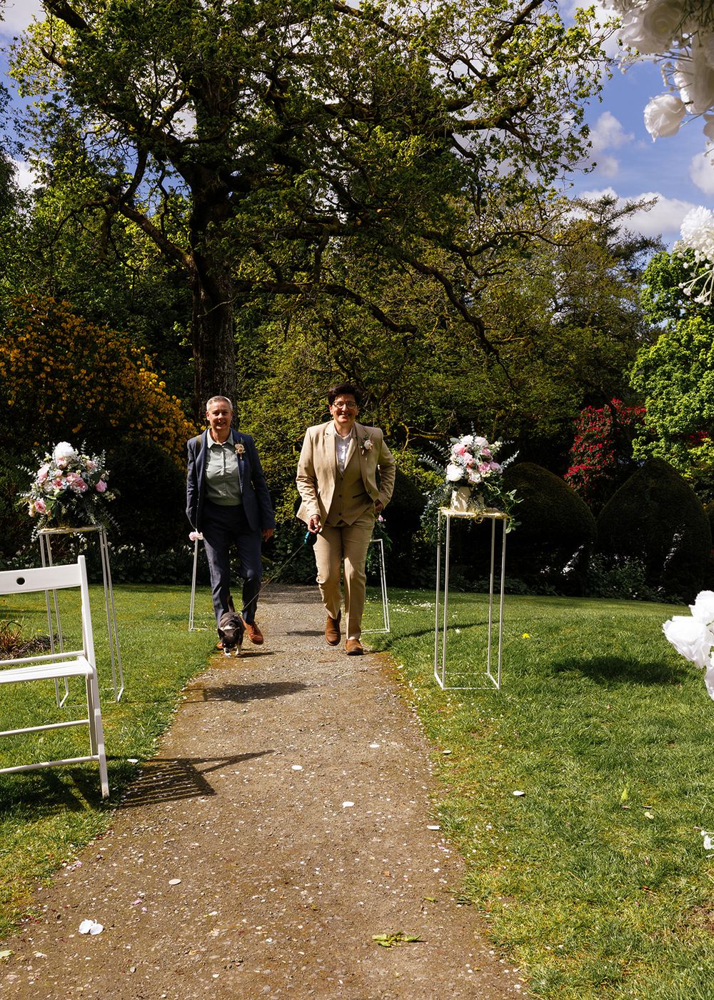 Two grooms walk down an outdoor wedding aisle lined with floral arrangements and white chairs in a lush garden.