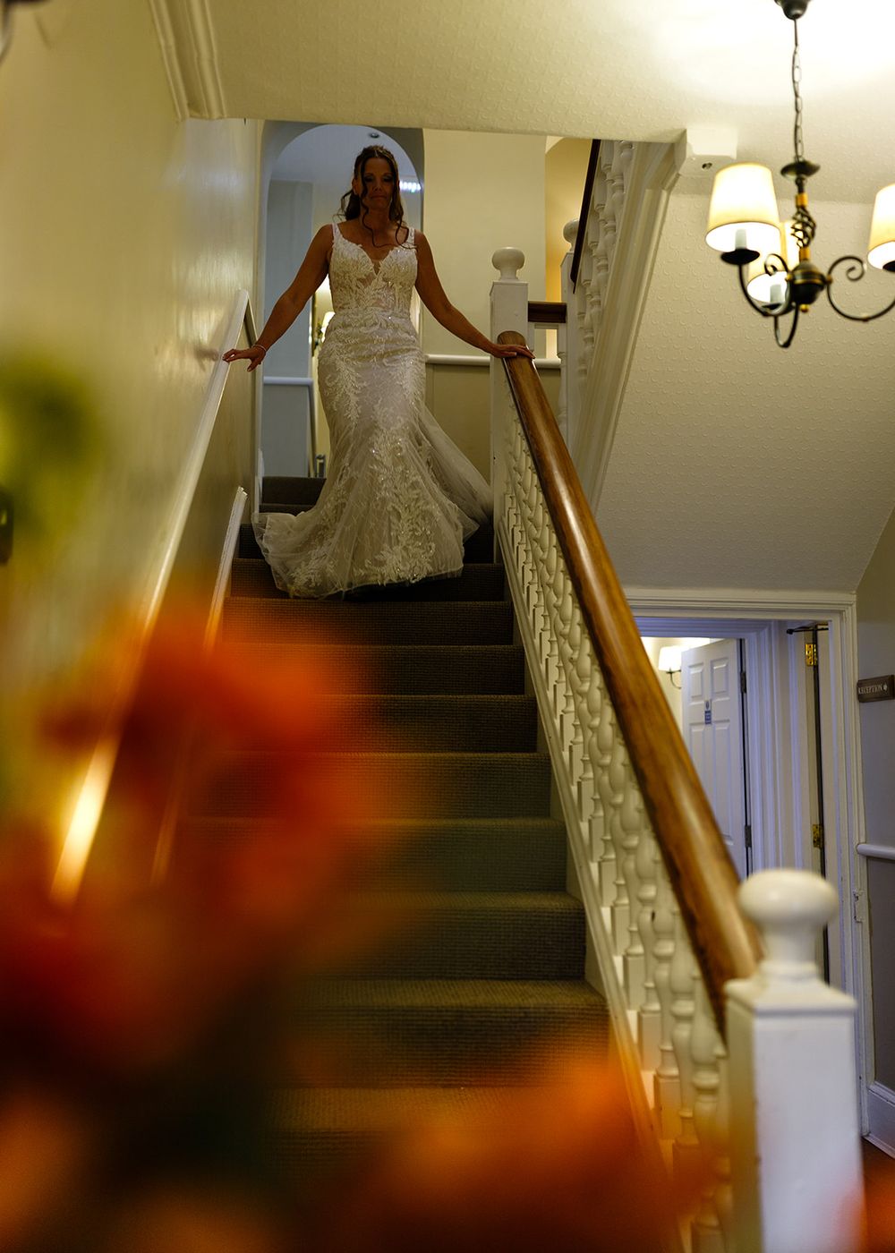 Bride in lace mermaid wedding dress descending staircase with chandelier and floral foreground.