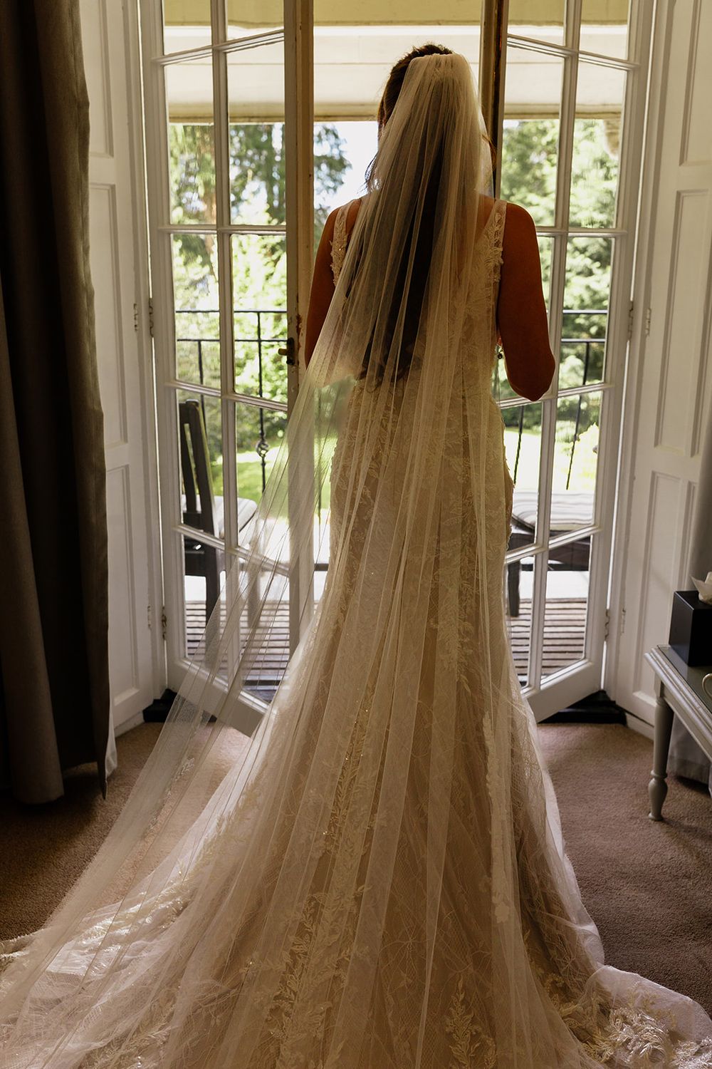 Bride in lace wedding gown and cathedral veil stands before open French doors with garden view.