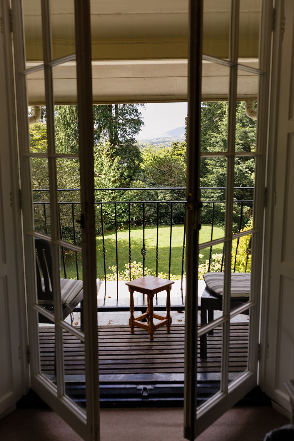 French doors open to a wooden balcony with a small stool, iron railings, and lush garden views beyond.