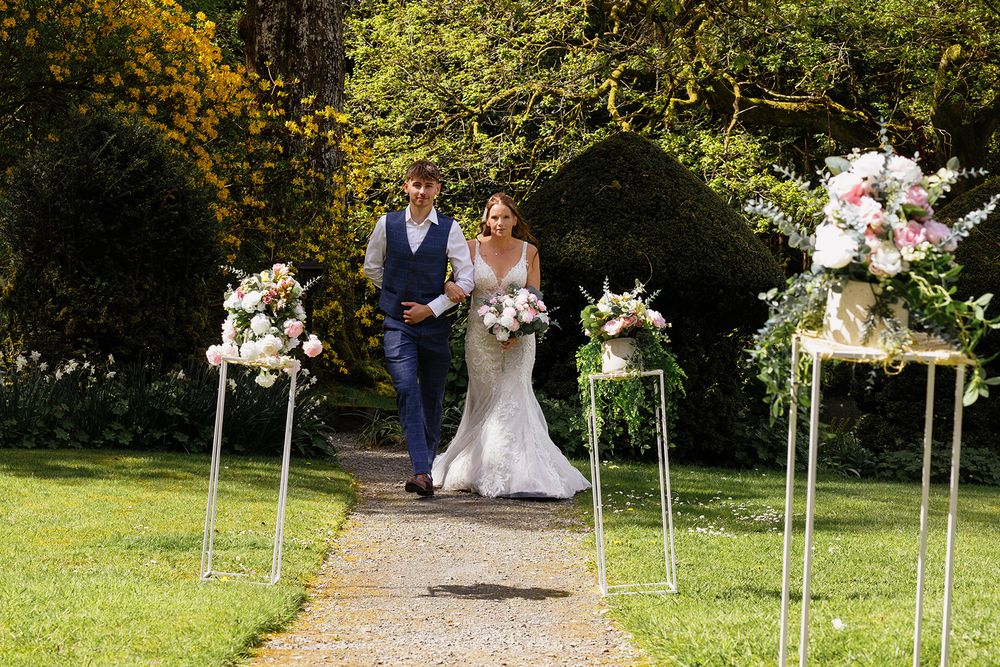 Bride and groom walking down outdoor aisle lined with floral arrangements on pedestals at a garden wedding ceremony.
