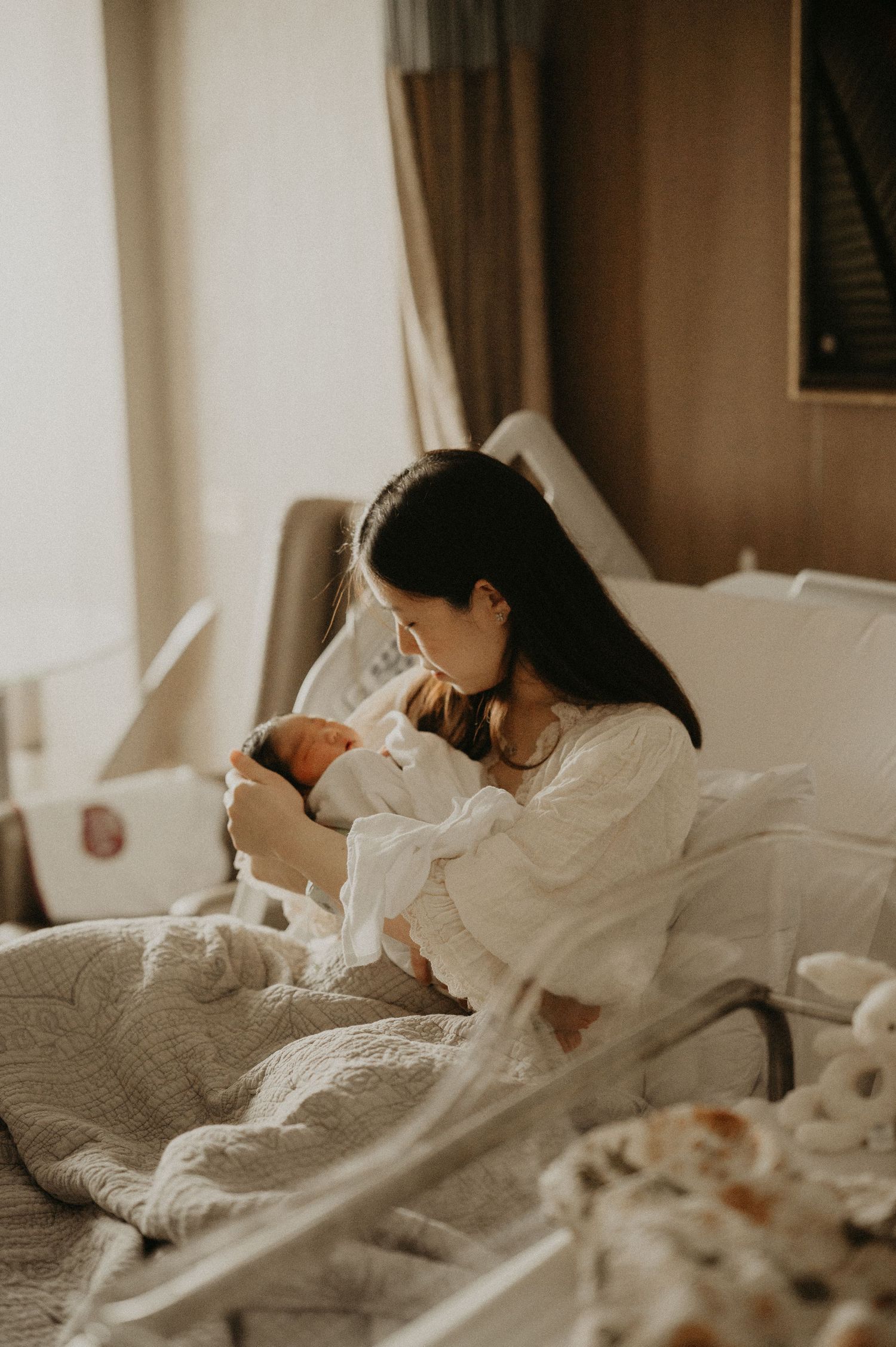 New mother with long dark hair cradles her swaddled newborn while resting in a hospital bed beside a clear bassinet in soft natural light.
