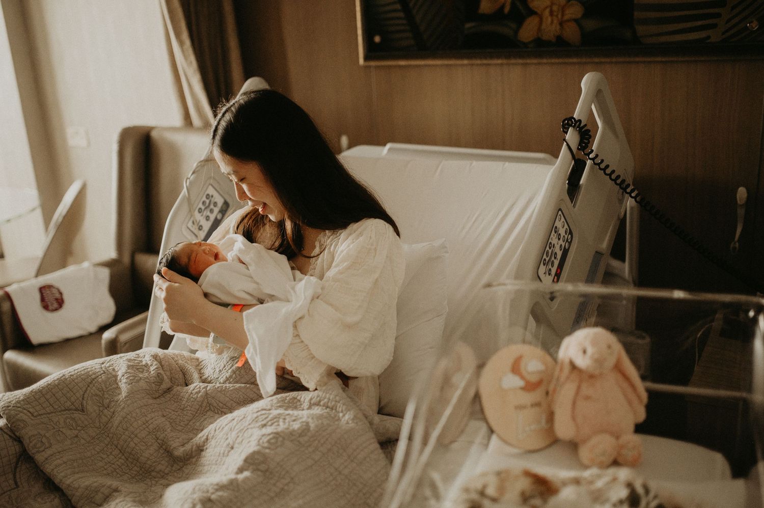 A mother in a white blouse gently kisses her sleeping newborn baby wrapped in a soft white blanket in a cozy, softly lit room.