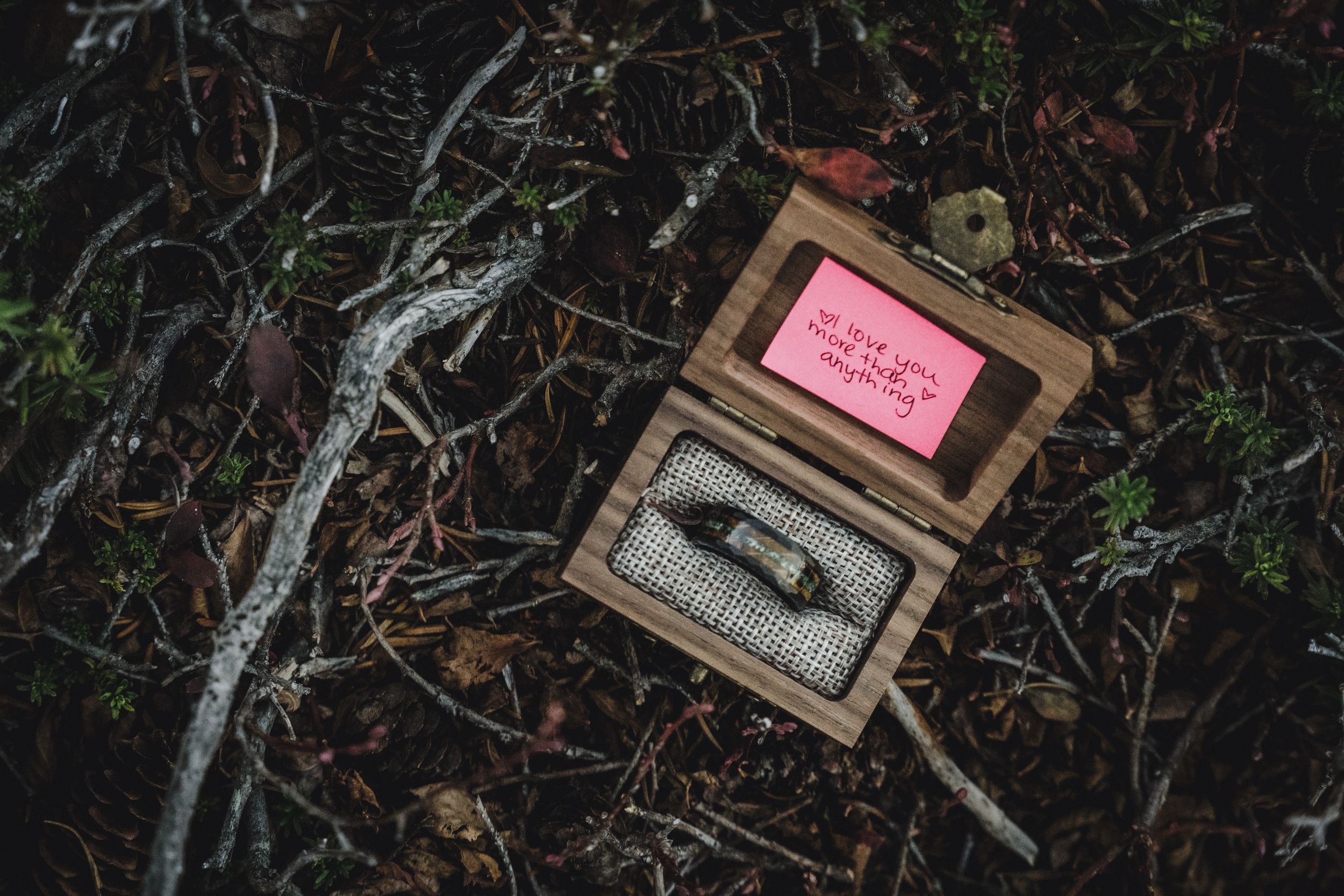 Open wooden ring box with a pink handwritten note inside the lid, resting on a forest floor of leaves, twigs and moss.