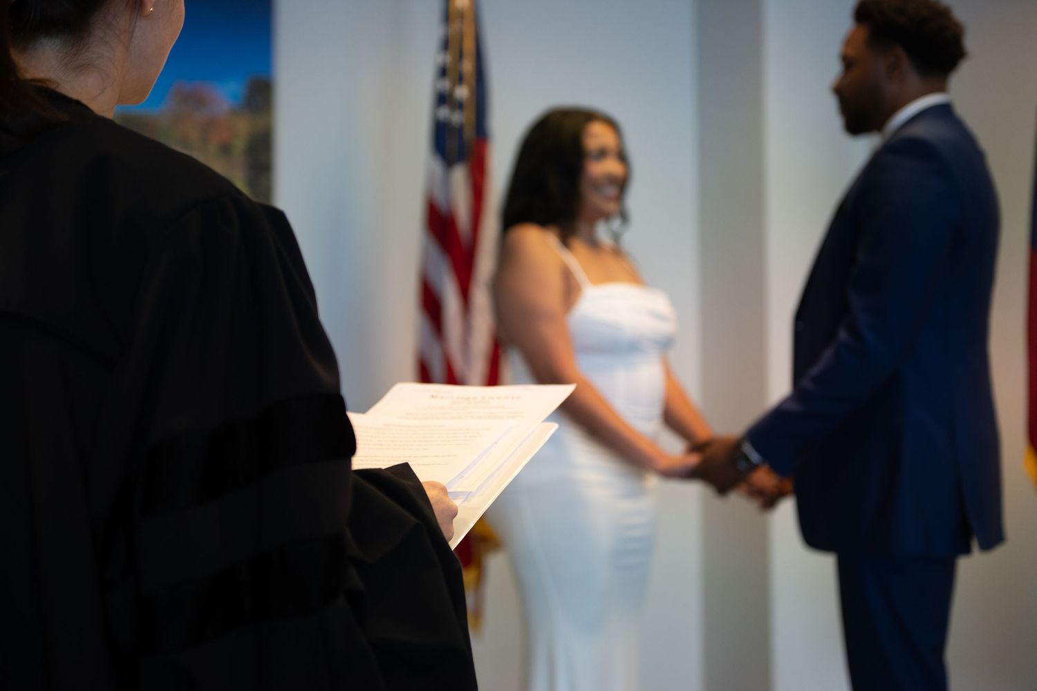 Bride in a white gown and groom in a navy suit hold hands during a wedding ceremony as an officiant reads, with an American flag behind.