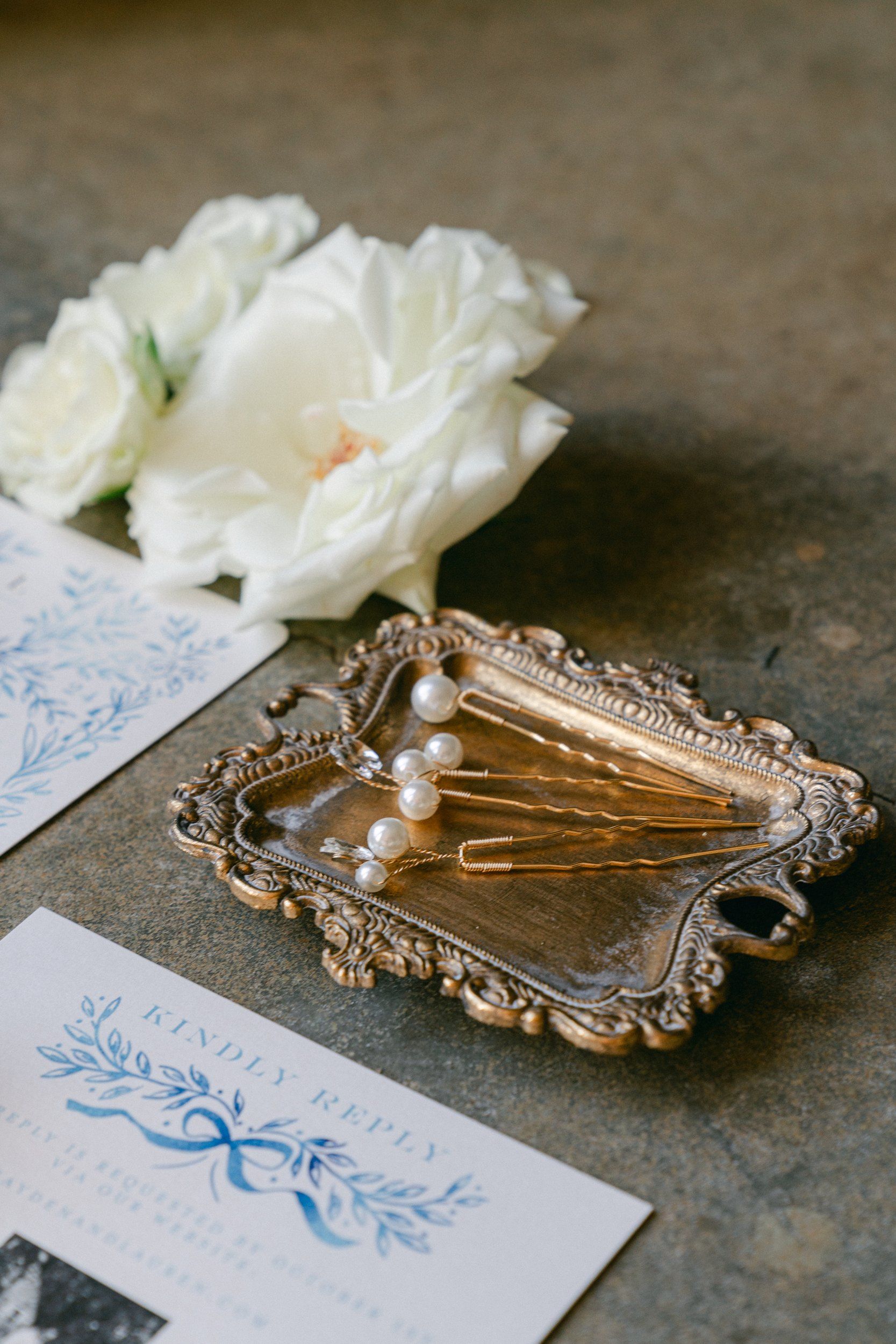 Pearl bridal hairpins on a gilded tray beside white roses and a blue watercolor wedding invitation