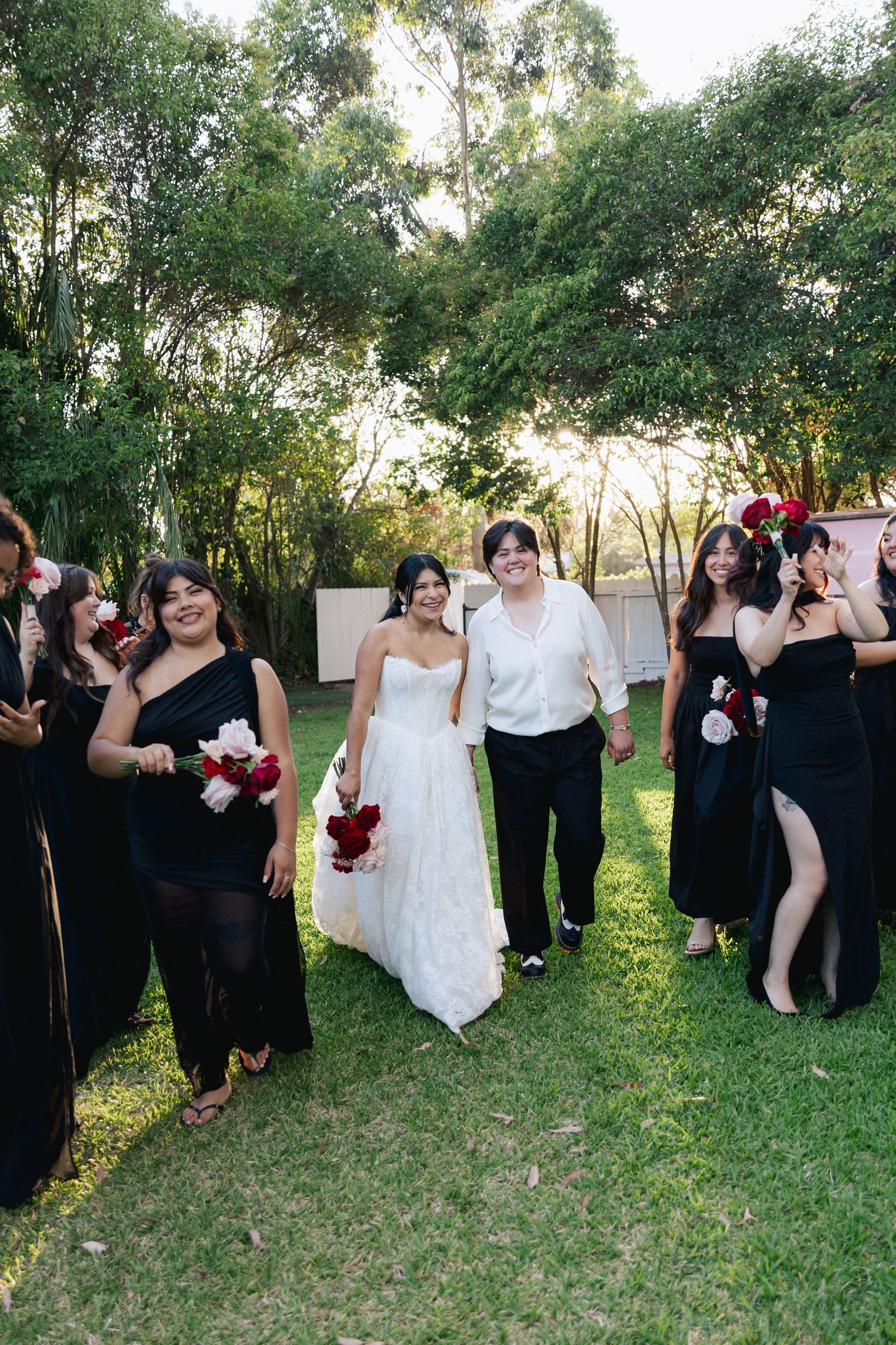 Bride in a white gown walks across a sunlit lawn with a partner in white shirt, flanked by bridesmaids in black dresses holding red bouquets