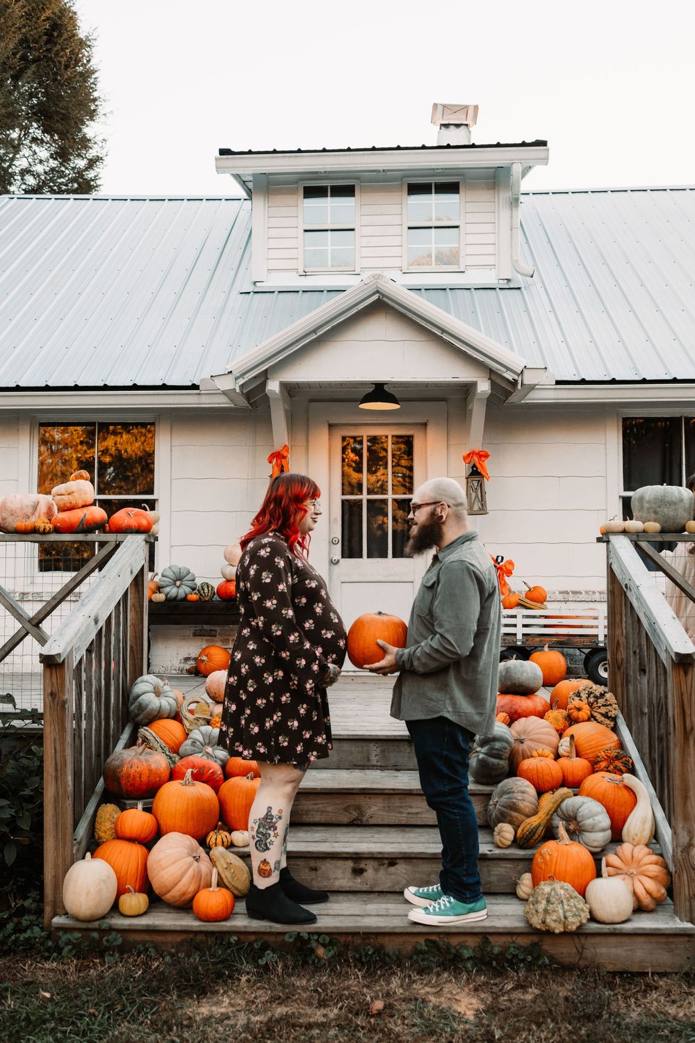 Couple holding a pumpkin on the steps of a white farmhouse porch surrounded by colorful autumn pumpkins and gourds.