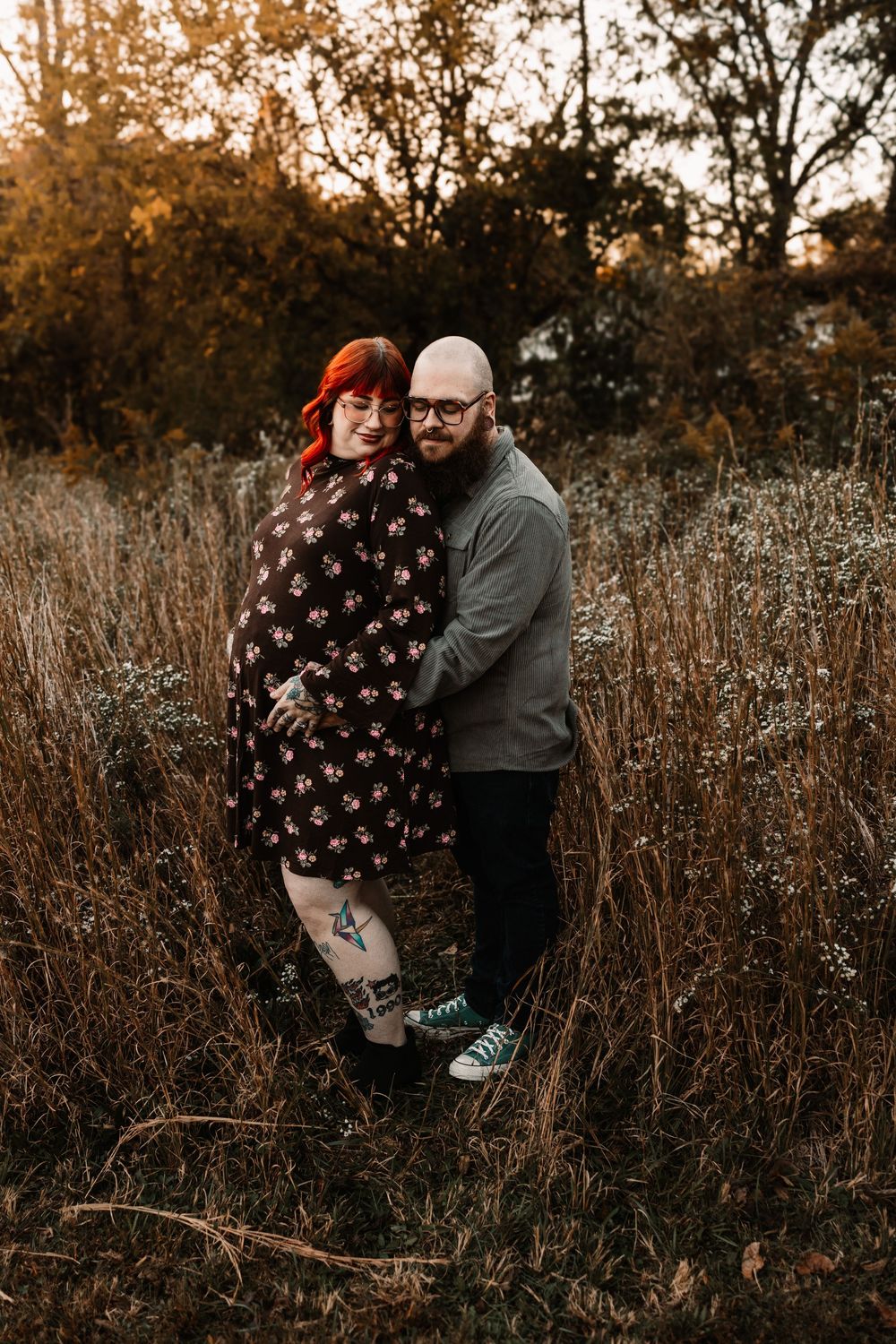 A smiling couple embraces in a tall grass field at sunset, surrounded by warm golden autumn trees with Nina Thomas Photography.