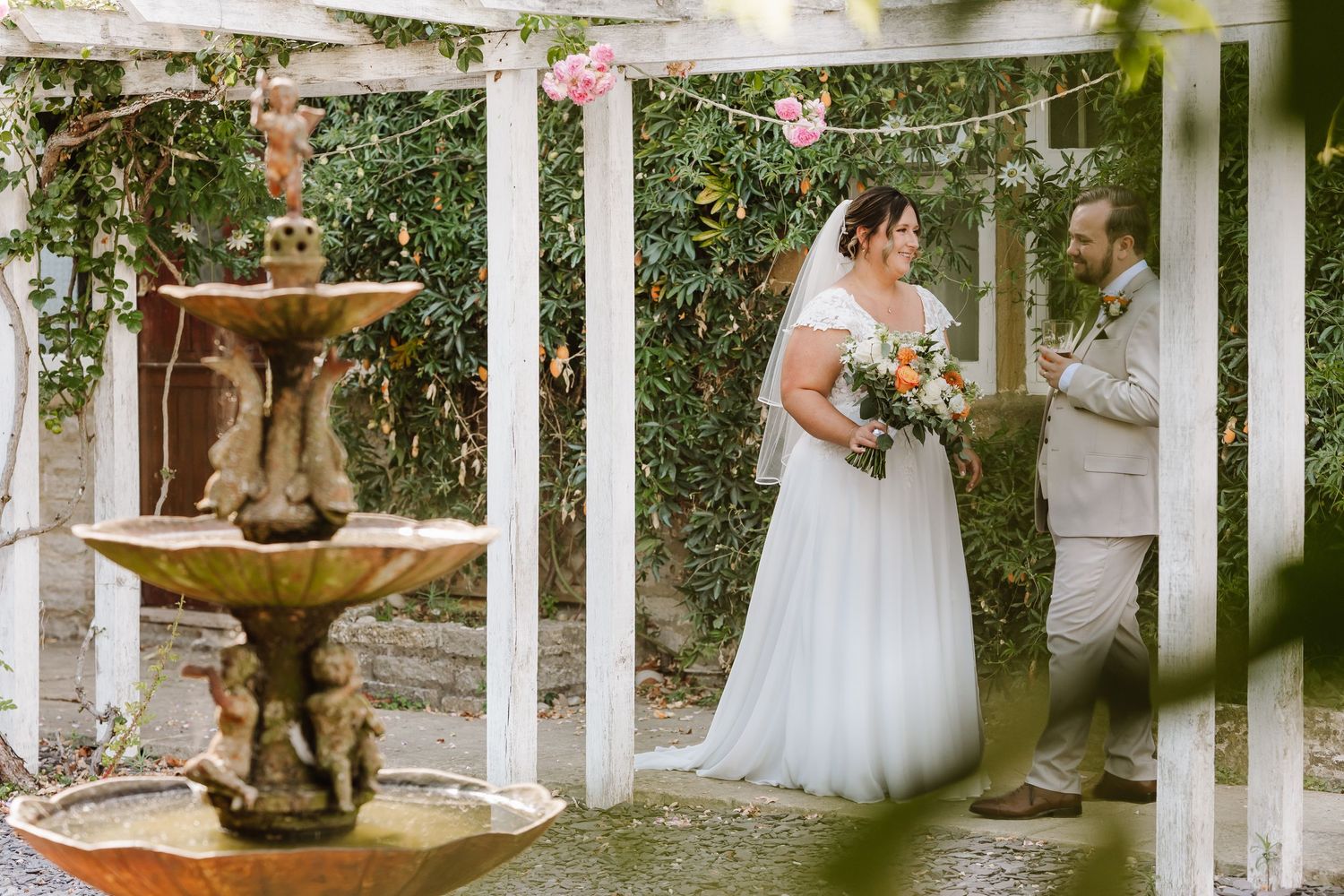 Bride in a white gown holding a bouquet stands with a groom beside a tiered stone fountain under a white pergola in a garden.