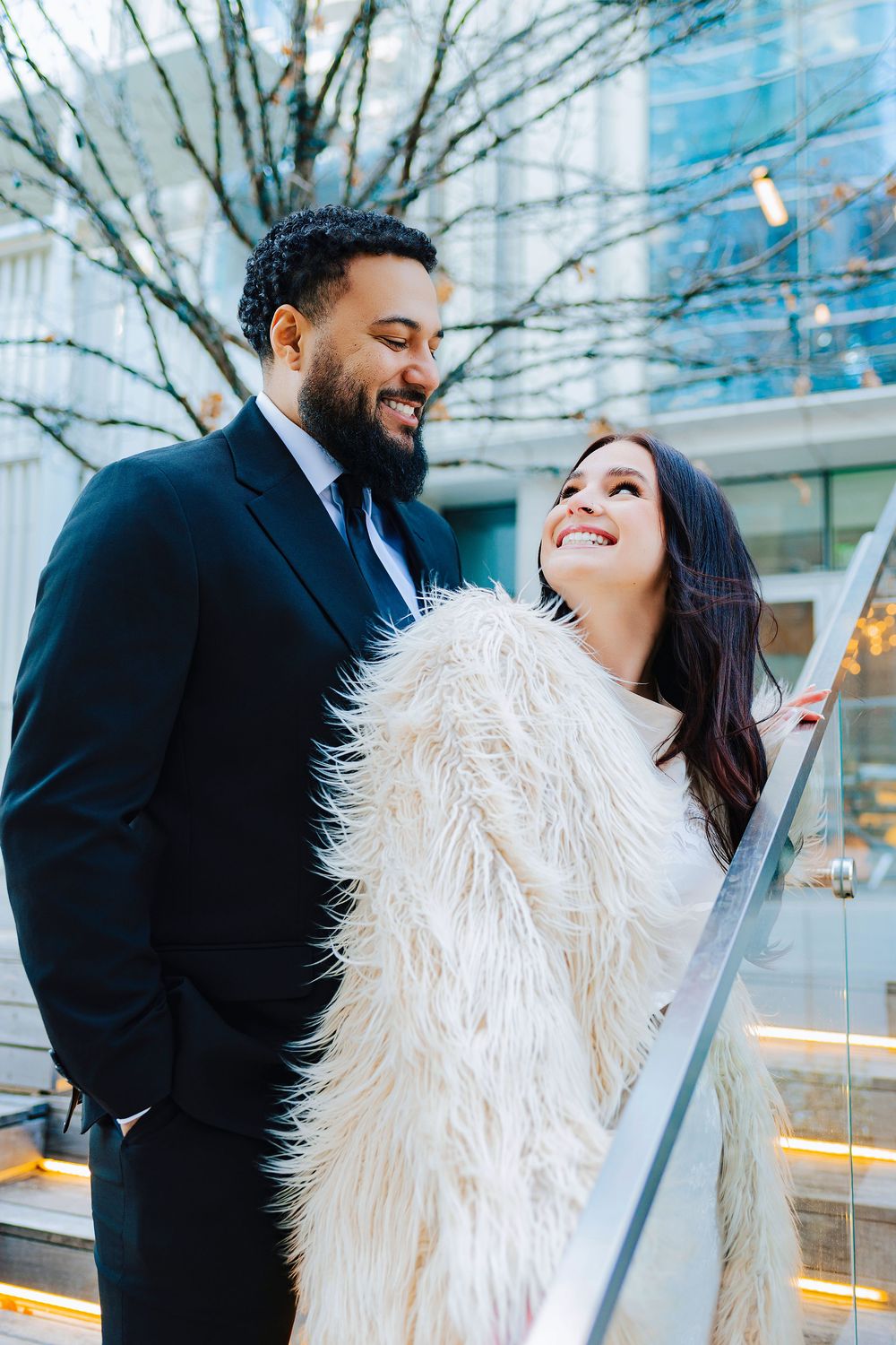 A smiling couple in elegant attire shares a romantic moment outdoors, the woman wearing a fluffy white coat.