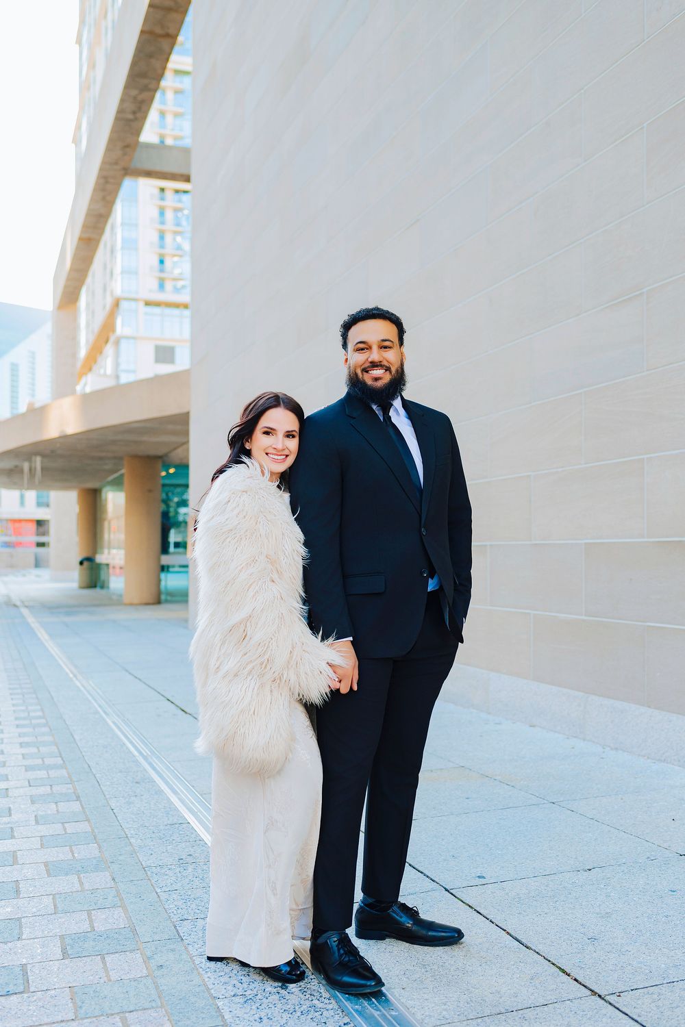 Couple posing outdoors, woman in white fur coat and man in navy suit, holding hands beside a modern building.