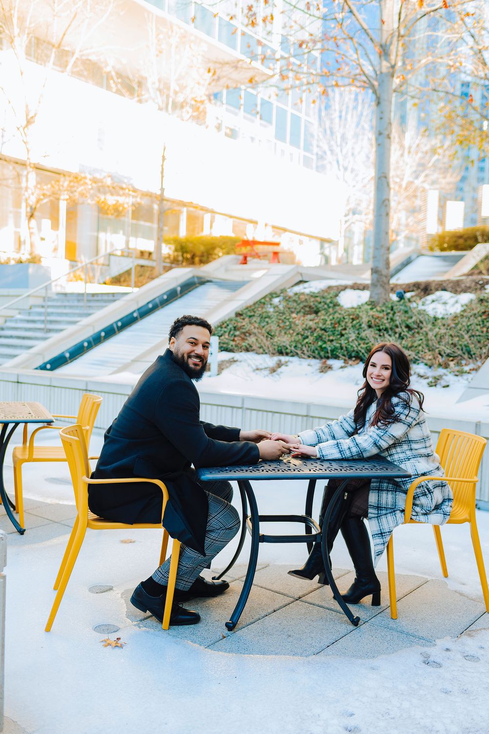 A smiling couple holds hands at an outdoor cafe table with yellow chairs, dressed in smart casual attire on a sunny winter day.