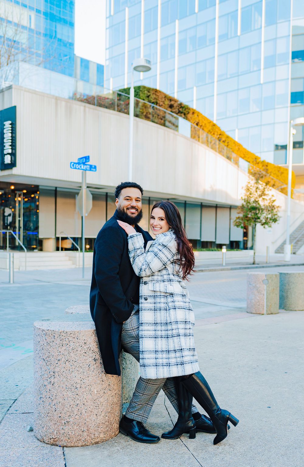A smiling couple poses together outdoors near a modern glass building, woman in plaid coat and man in black suit.