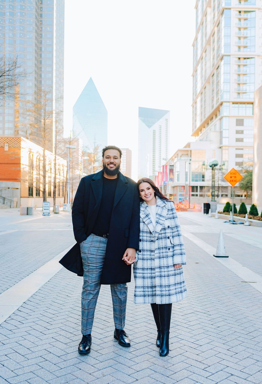 A couple holding hands and smiling in a city plaza, wearing stylish plaid coats with modern skyscrapers in the background.