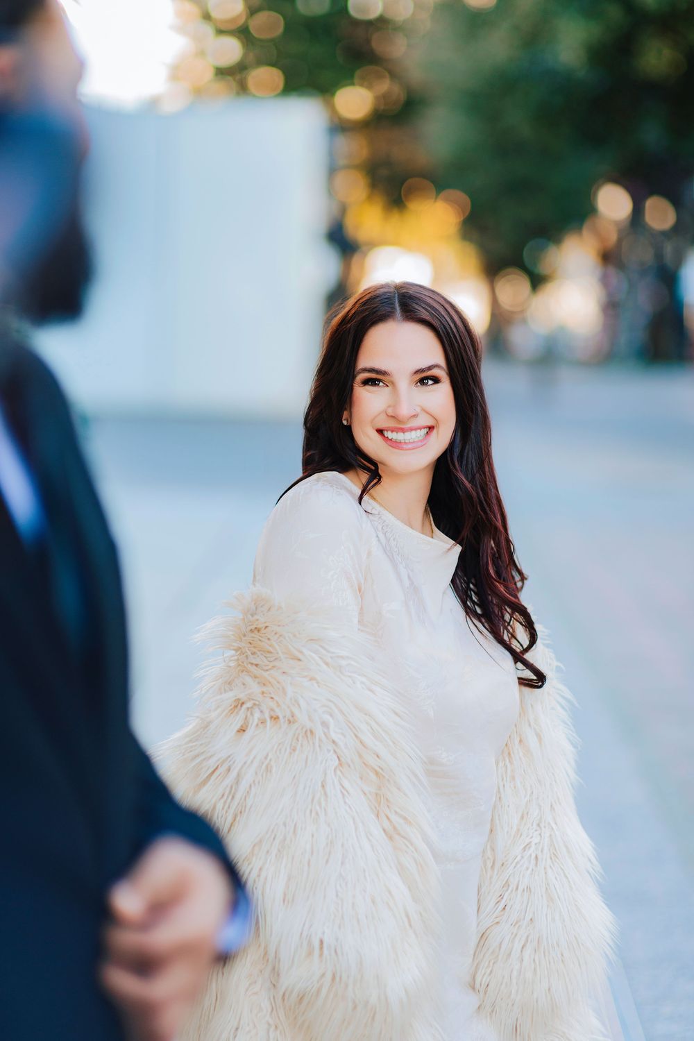 Smiling brunette woman in white faux fur coat holding hands outdoors in soft bokeh sunlight.