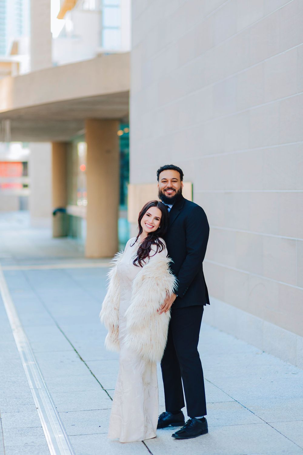 Elegant couple posing outdoors, woman in white fur coat and man in black suit, smiling against a modern building.