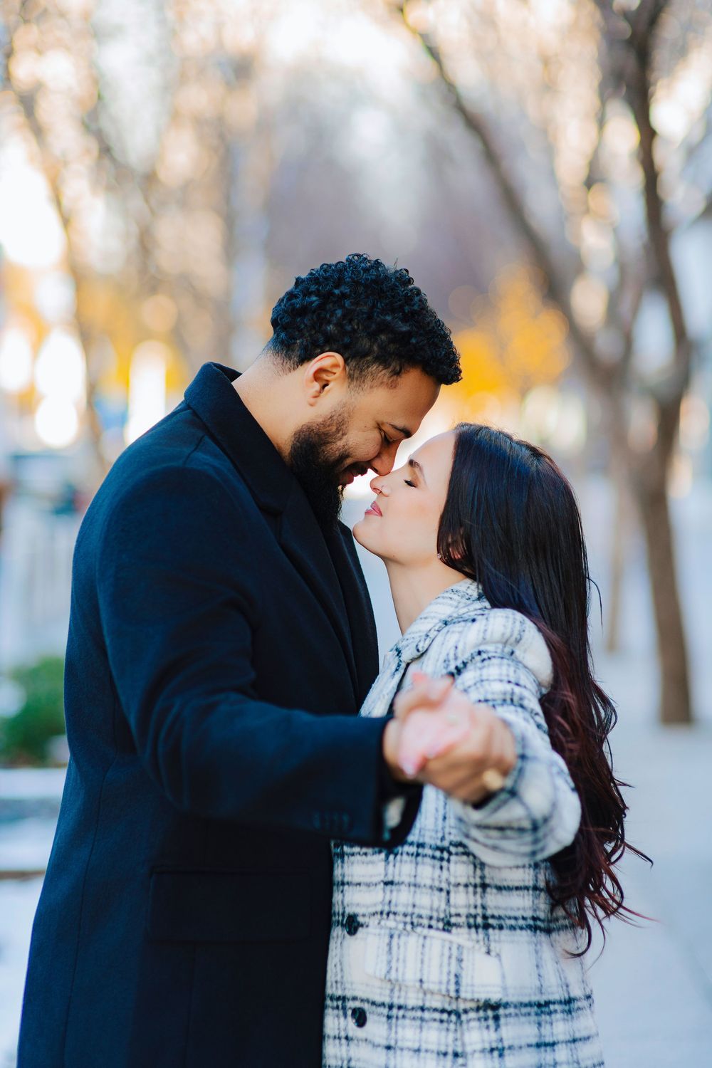 A couple in winter coats share a romantic moment outdoors, leaning in for a kiss among autumn trees.