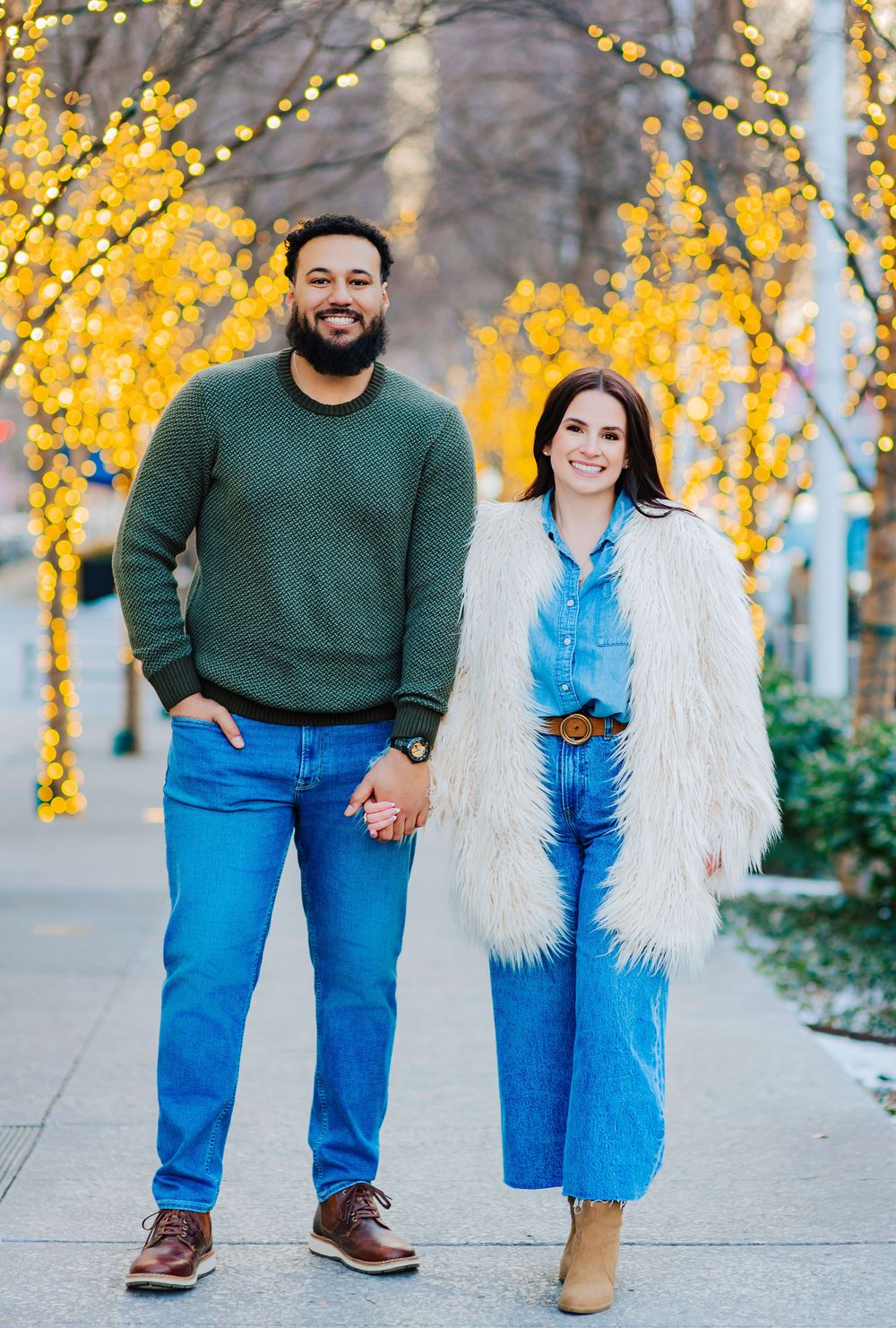 Couple holding hands smiling outdoors with yellow string lights on trees in winter, man in green sweater, woman in white fur coat.