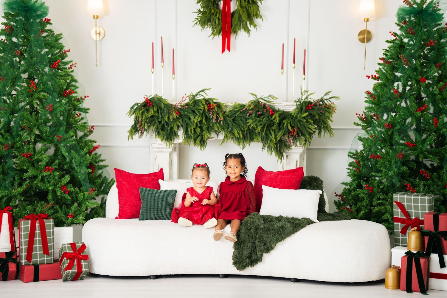 Two young girls in red dresses sit on a white sofa surrounded by Christmas trees and festive holiday decorations.