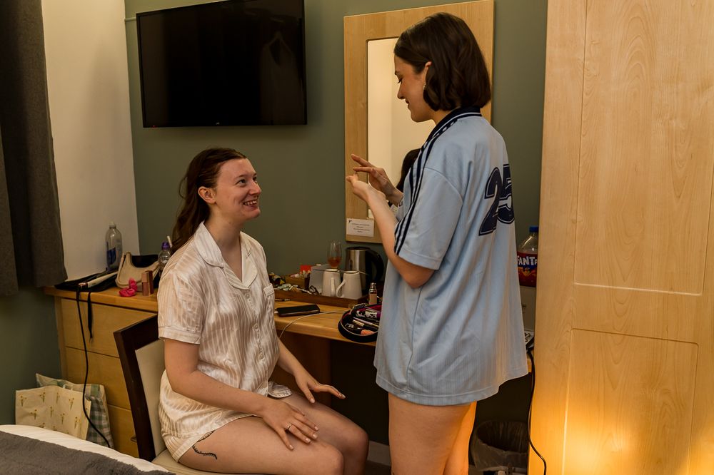 Two women chatting in a hotel room, one seated at a vanity desk, the other standing in a blue jersey.