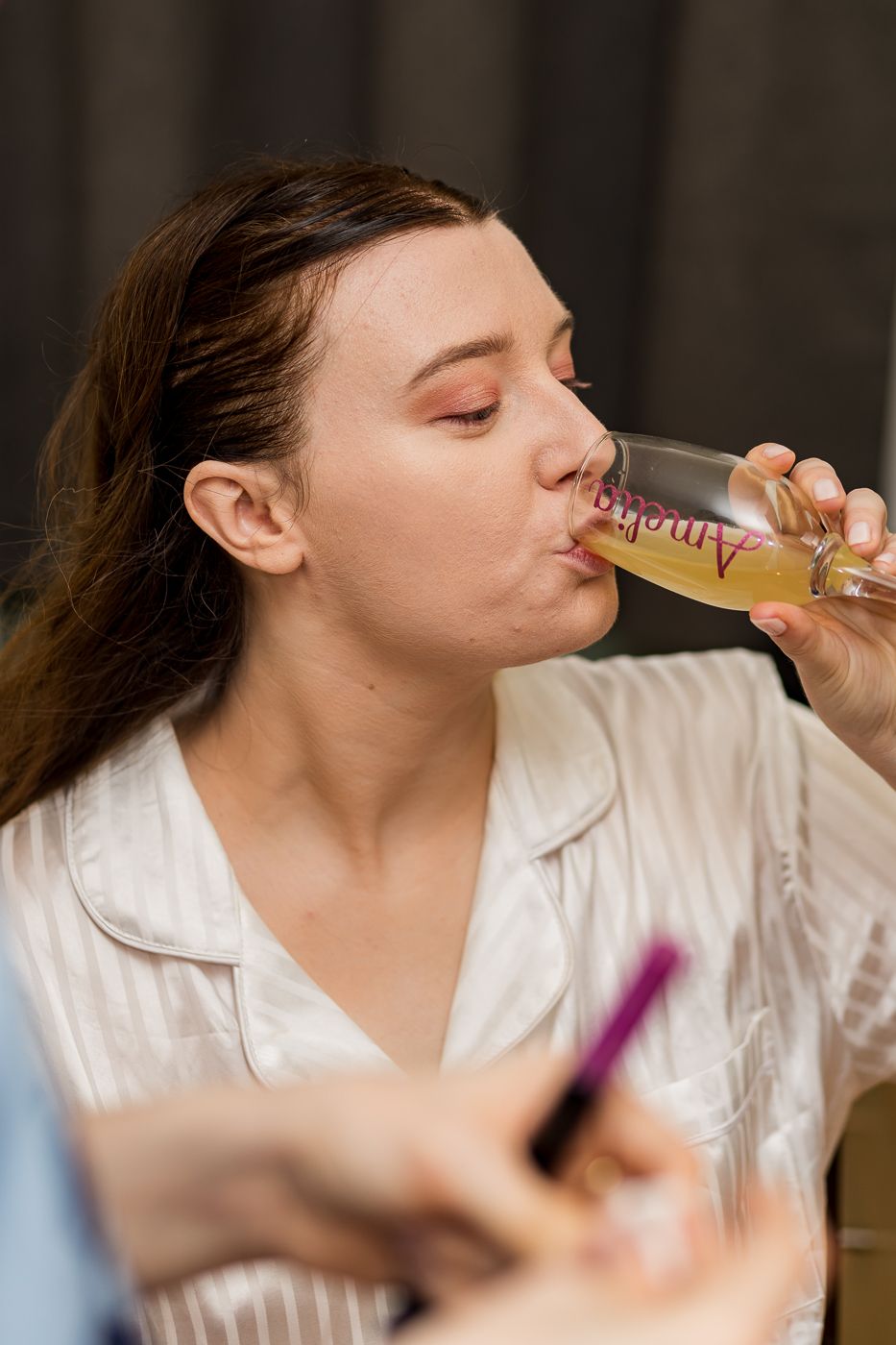 Woman in white striped pajamas sipping champagne from a personalized glass during a bridal getting-ready session.