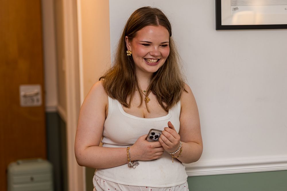 Young woman smiling and holding a phone, wearing a white tank top and gold jewelry indoors.