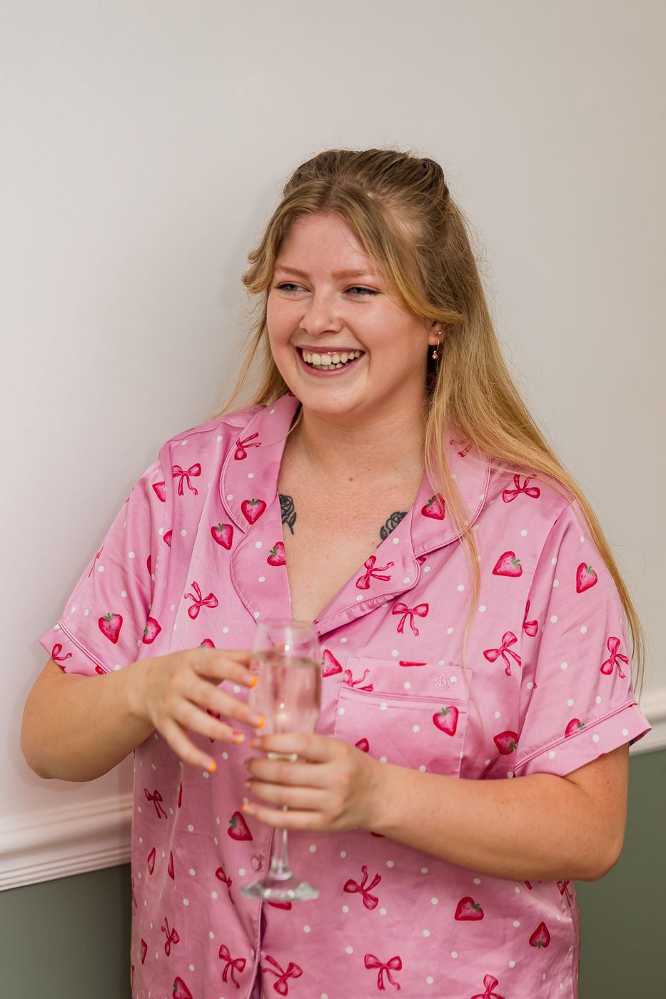 Smiling woman in pink heart and bow print pajamas holding a champagne glass against a white wall.