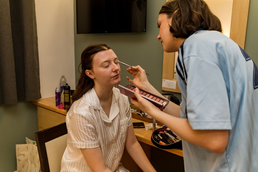 A makeup artist applies eyeshadow to a young woman sitting at a vanity mirror in a softly lit room.