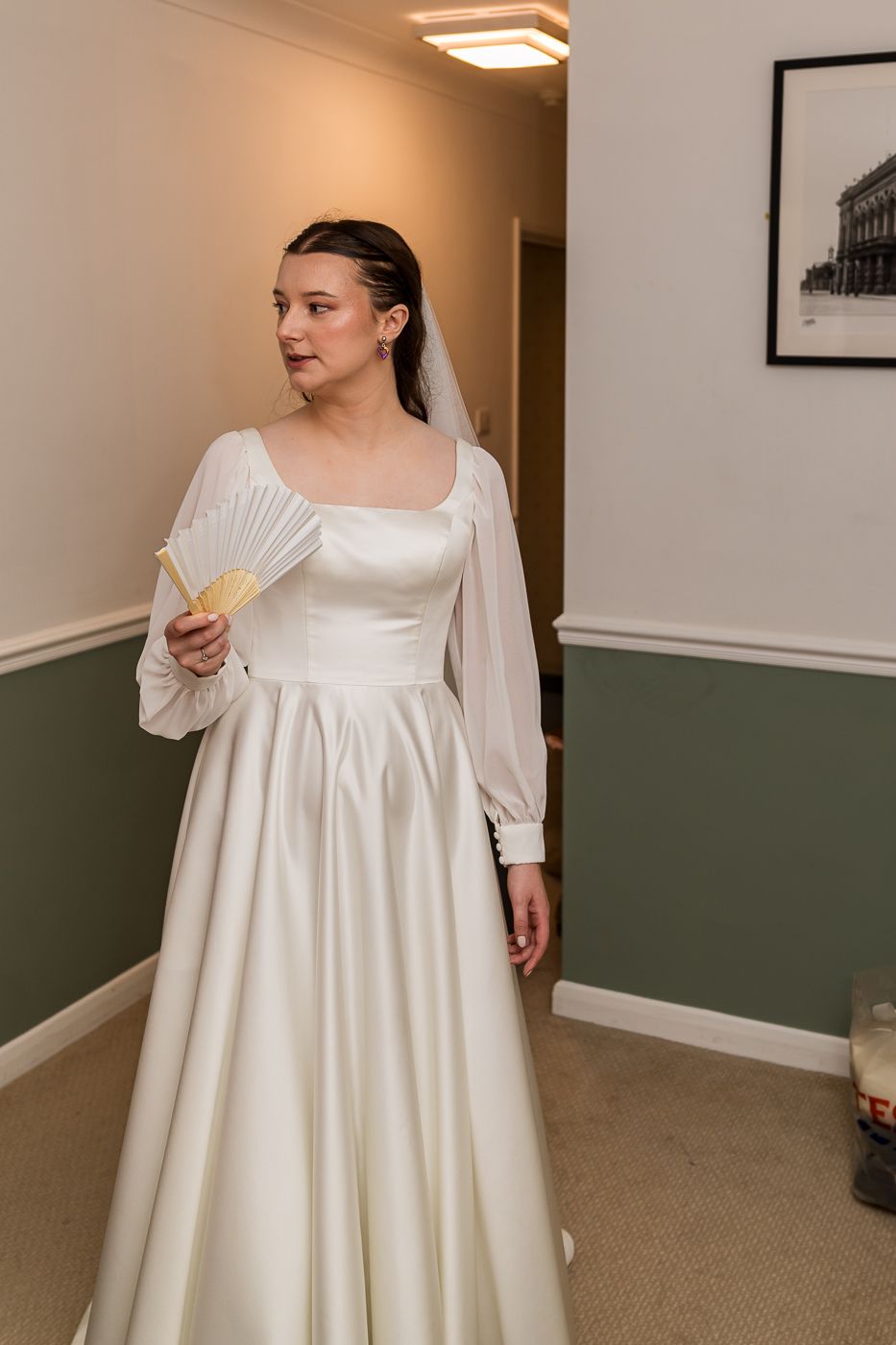 Bride in elegant white satin gown with sheer sleeves holding a fan, wearing veil in a softly lit room.