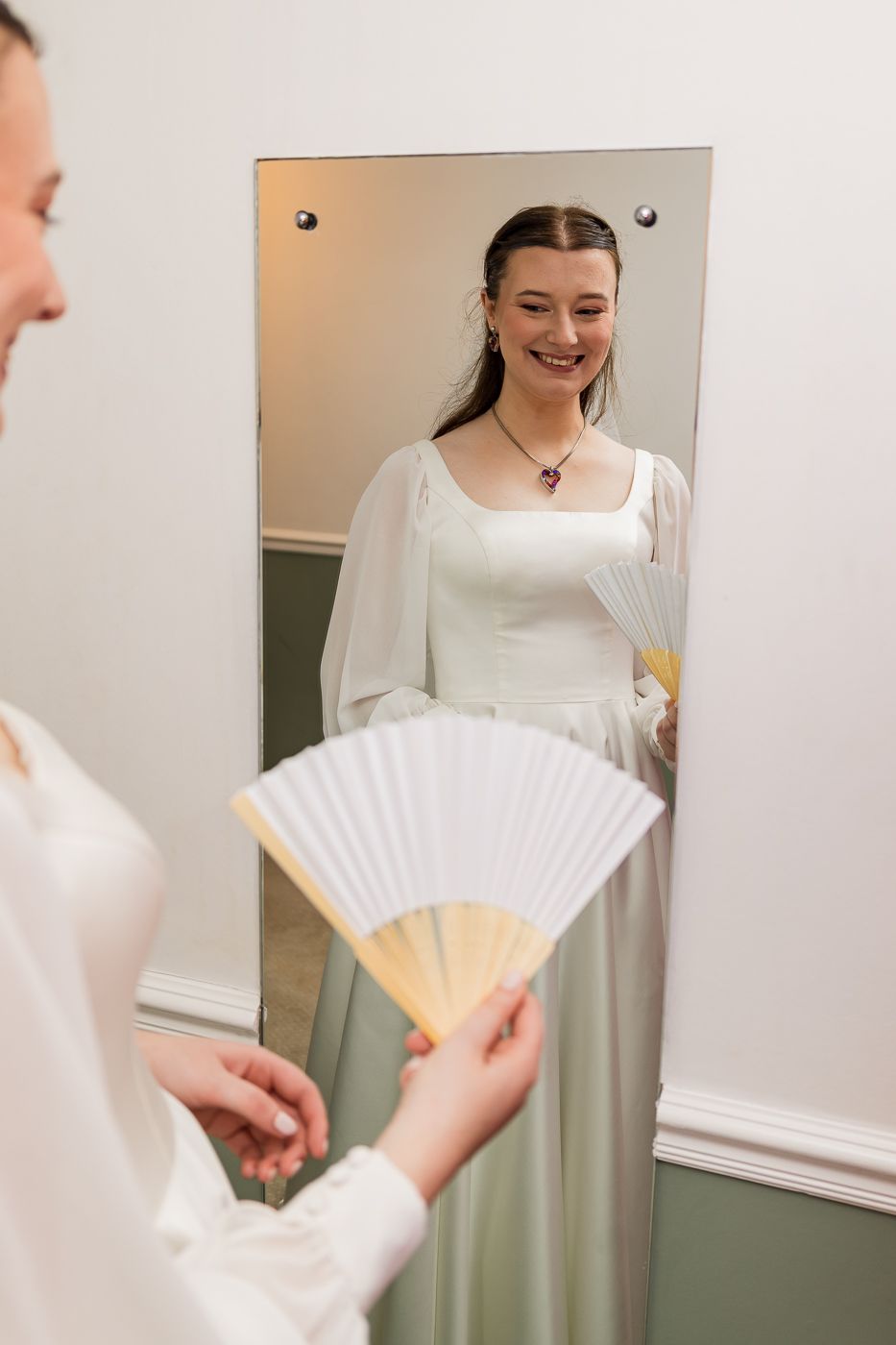 Smiling bride in white gown holding a decorative fan, reflected in a full-length mirror.