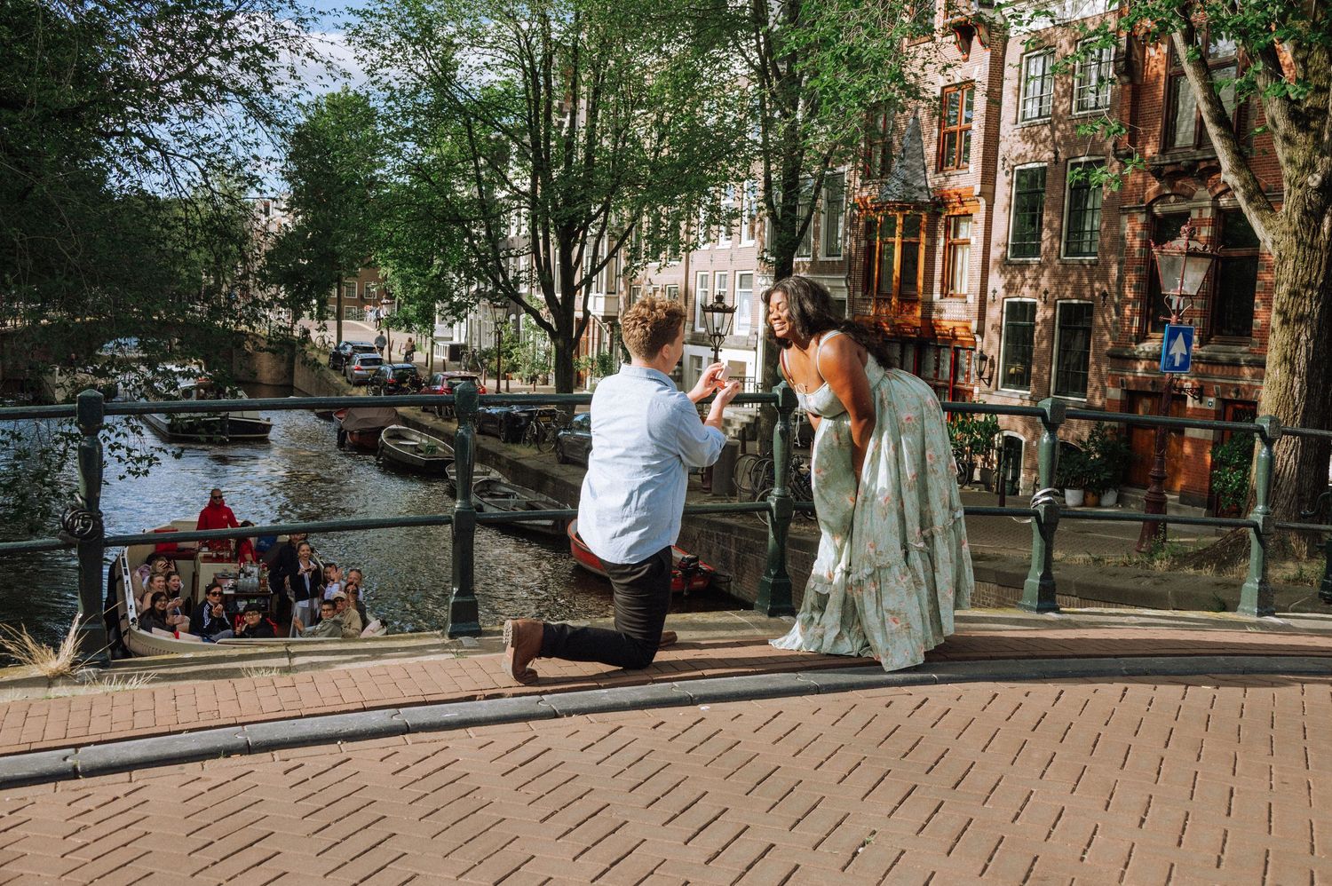 Man kneeling on one knee proposing to a smiling woman in a long white dress on a canal bridge Amsterdam.
