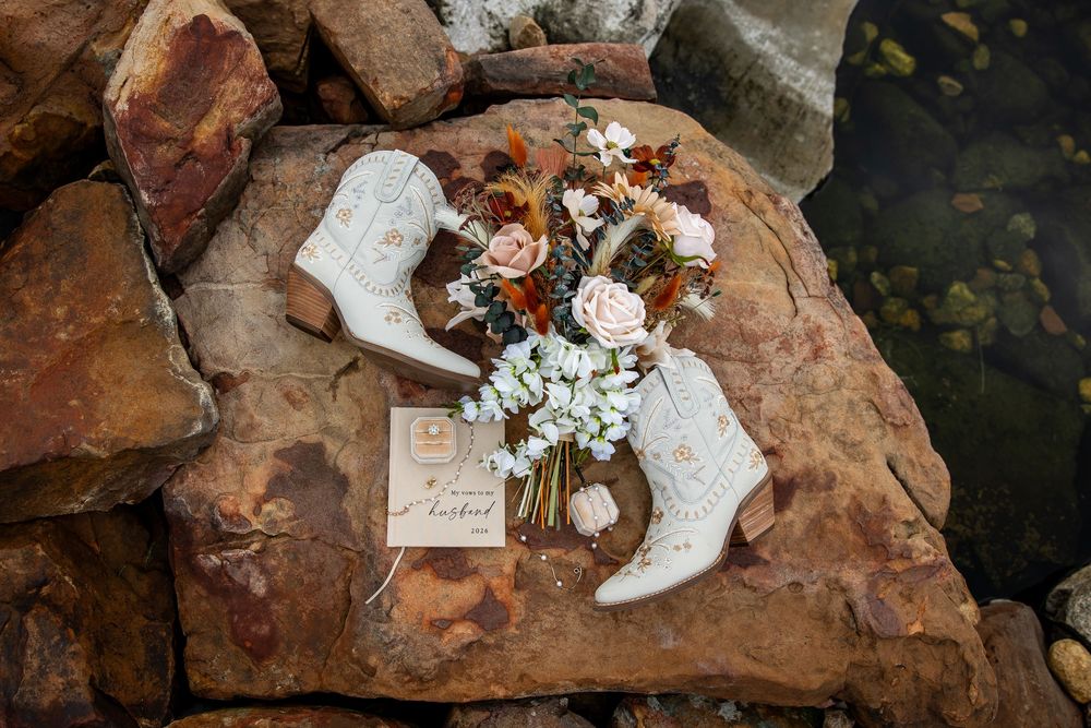 White cowboy boots and a boho bridal bouquet of peach roses and wildflowers rest on a red rock beside a letter, with water nearby.