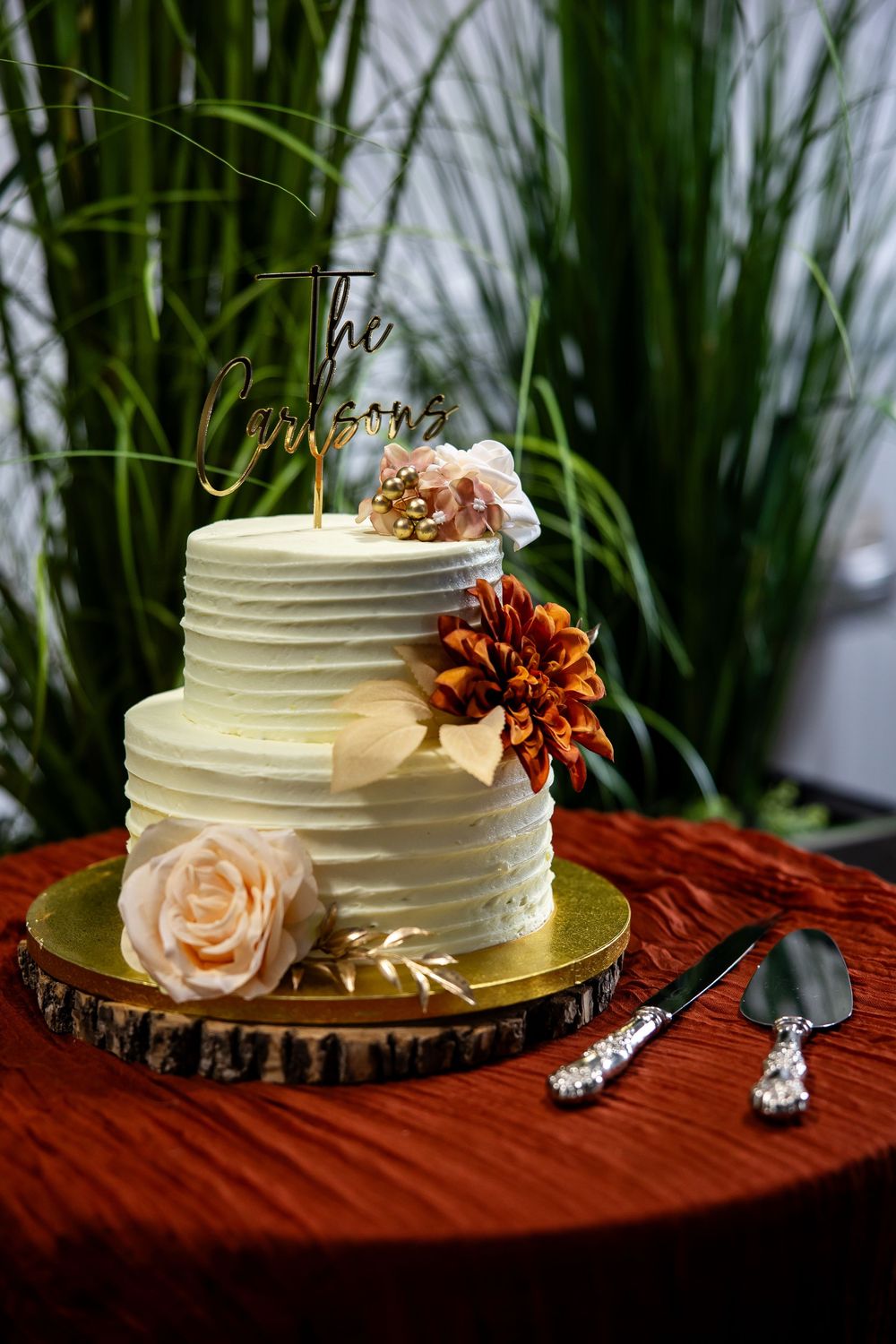Two-tier buttercream wedding cake with a gold topper, blush rose and rust dahlia flowers, on a wood slice stand with cake server set.