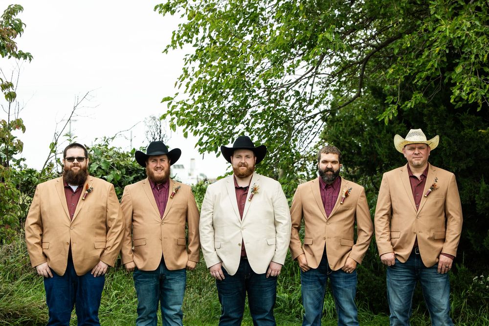 Five bearded groomsmen in tan blazers, burgundy shirts and jeans, some wearing cowboy hats, stand in a row outdoors in front of green trees.