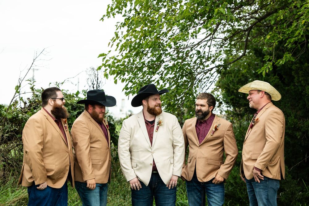 Six bearded men in tan and cream western blazers and cowboy hats stand together laughing outdoors in front of green leafy trees.